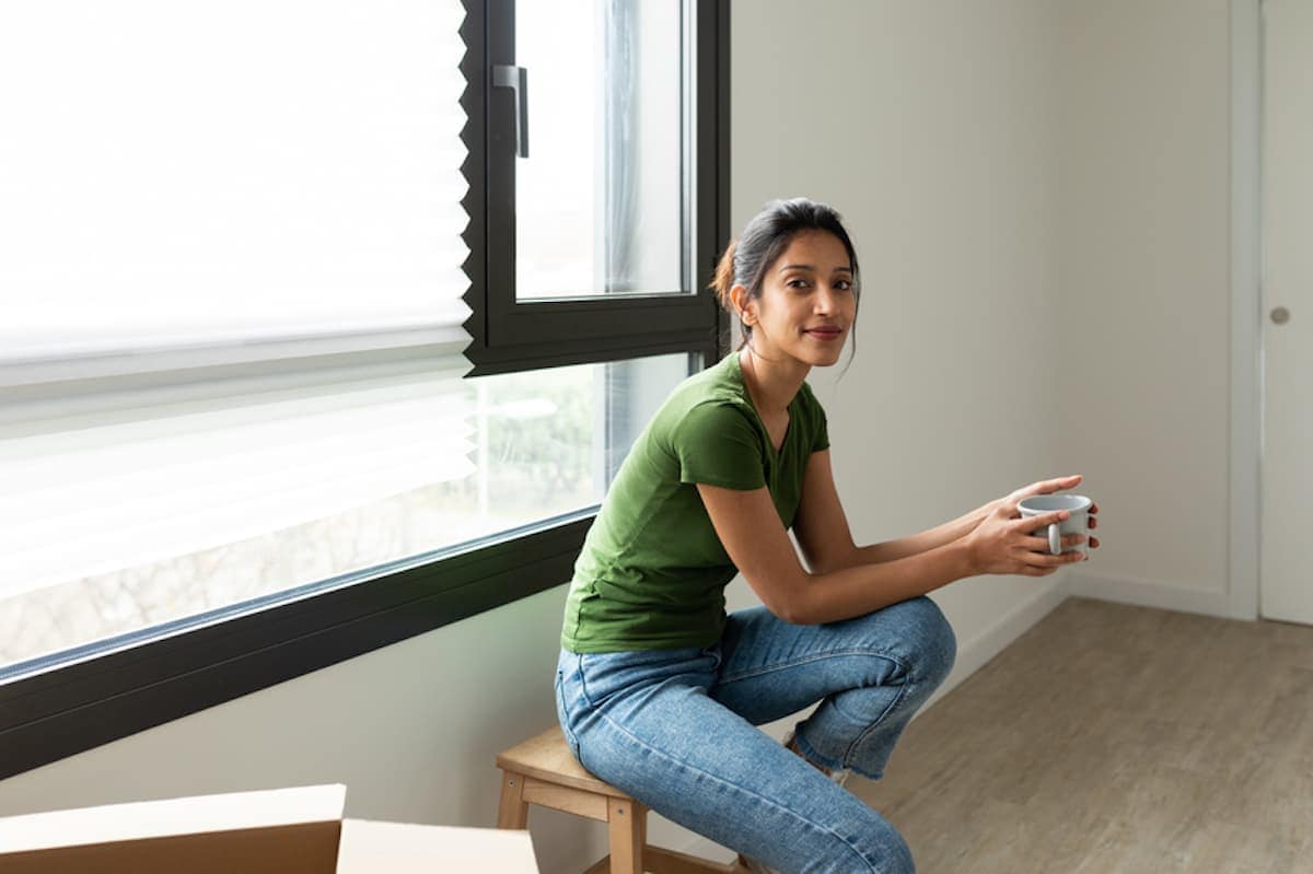 A young woman sits on a stool next to a moving box in an empty room.